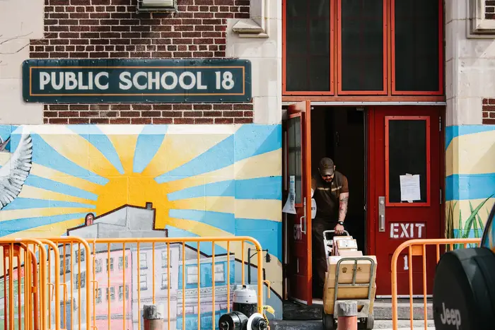 A UPS delivery man in a brown uniform pulls a dolly with boxes through the red front doors of a School, which has a colorful mural on the outside